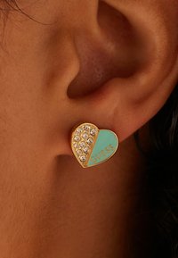 Boucles d'oreilles en forme de cœur en or, présentant de l'émail turquoise d'un côté et des strass clairs de l'autre, avec "GUESS" embossé en orange.