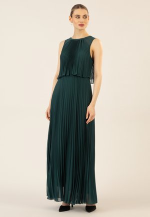 Faltenmaxikleid in tiefgrün, mit ärmellosem Design und geschichteter Brustpartie. Der Stoff hat eine glatte Textur und wird mit angespitzten schwarzen Absätzen kombiniert.