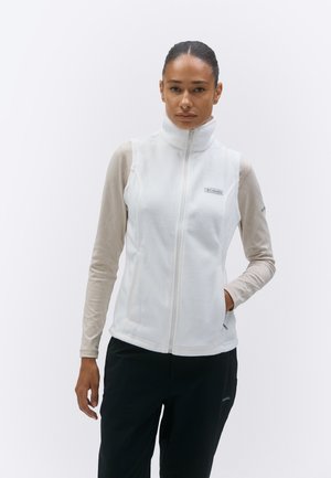 Γυναίκα με λευκό φλις γιλέκο με φερμουάρ πάνω από μπεζ μακρυμάνικη μπλούζα και μαύρο παντελόνι, στέκεται με το ένα χέρι στην τσέπη μπροστά σε απλό φόντο.