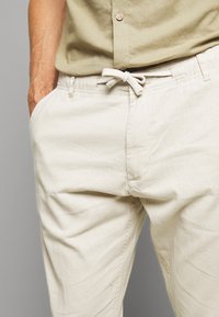 Pantalones de algodón de color beige claro con cintura elástica, cierre con cordón y bolsillos laterales; textura suave con detalles de costura mínimos.