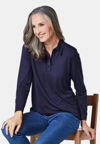 Marineblaues Langarm-Polohemd mit einer Drei-Knopf-Leiste und geripptem Kragen, kombiniert mit blauen Jeans, auf einem Holzstuhl sitzend.