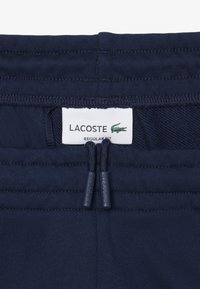 Pantaloni Lacoste blu navy con cintura elastica, dotati di due tasche con zip, etichetta con marchio e dettagli in tessuto testurizzato.