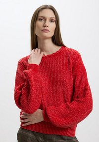 Maglione rosso a coste con maniche larghe e scollatura tonda. Tessuto testurizzato e vestibilità morbida, indossato sopra pantaloni a quadri.
