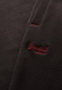 Ciemna tkanina z czerwonym wyszywanym logo "SuperDry" oraz japońskimi znakami blisko kieszeni z czerwonym przeszyciem.