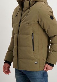 Olivgrön pufferjacka med huva, som har en framdragbar dragkedja, elastiska ärmslut och fickor med dragkedja. Inkluderar en logotyp på ärmen.