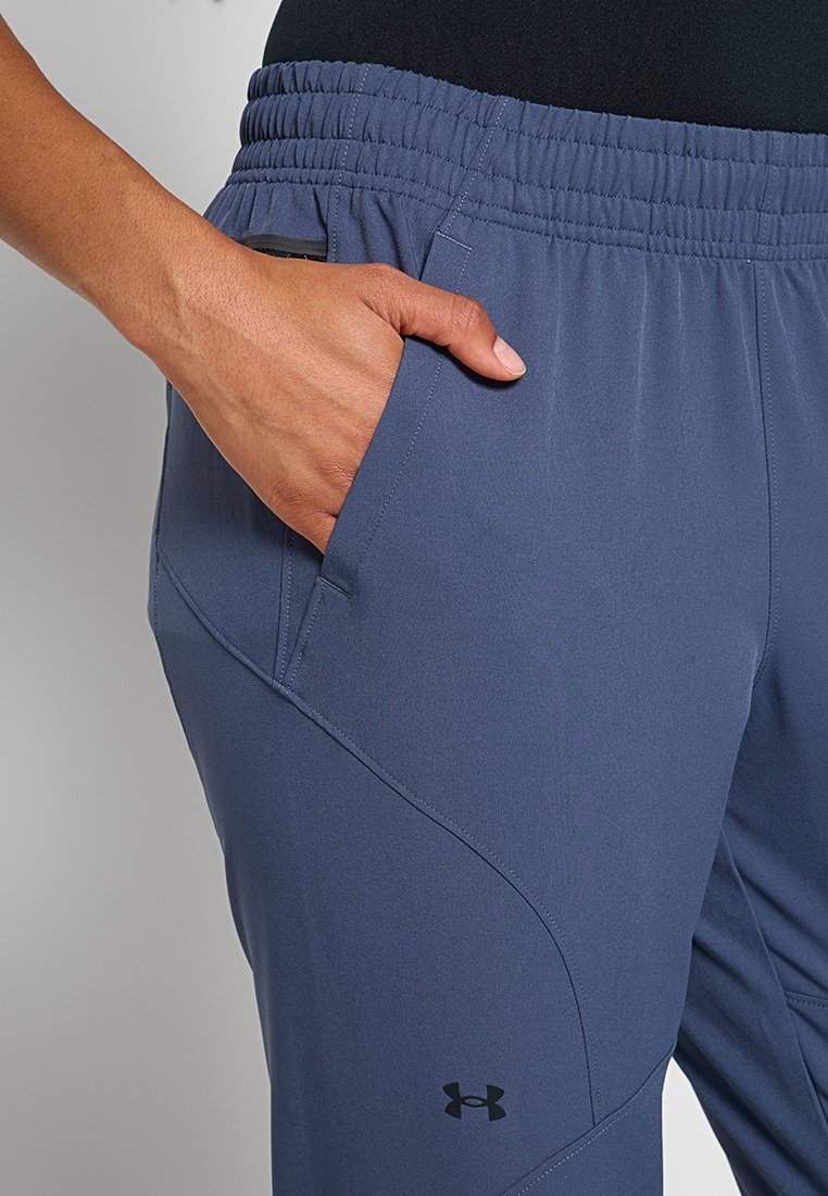 Main dans la poche d'un pantalon de sport bleu avec une taille élastique et le logo noir d'Under Armour sur la cuisse.