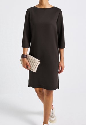 Vrouw draagt een zwarte jurk tot op de knie met driekwartmouwen, houdt een beige geweven clutch vast en draagt witte sneakers.