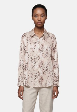 Woman wearing a light beige and brown spotted long-sleeve button-up blouse with beige pants standing against a plain background.