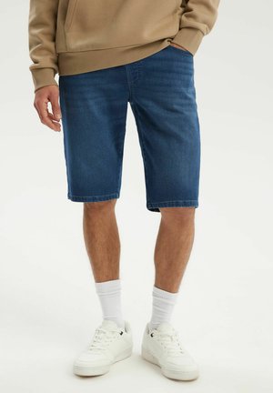 Mężczyzna w niebieskich jeansowych szortach, beżowej bluzie, białych skarpetach i białych trampkach, stoi z jedną ręką w kieszeni.