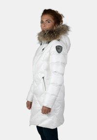 Giacca bianca imbottita con cappuccio in pelliccia sintetica removibile, texture a quadri di diamante, tasche laterali con zipper e una toppa del logo sulla spalla.