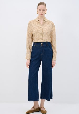 Camicia beige a fantasia con bottoni, abbinata a pantaloni in denim a gamba larga di colore blu scuro a vita alta. Le scarpe ballerine in camoscio marrone completano l'outfit.