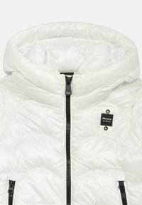 Casaco puffer branco com colarinho alto, apresentando um fecho éclair frontal e padrão acolchoado. Etiqueta de marca cosida no lado esquerdo.