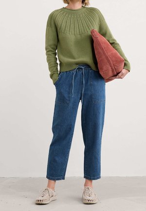 Persona con suéter verde oliva, pantalones vaqueros azules holgados, zapatos tejidos beige y sosteniendo un bolso grande de ante color óxido.