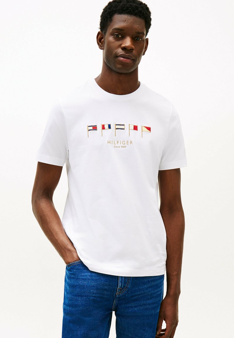 T-shirt bianco in cotone con collo rotondo, dotato di grafiche di bandiere multicolori e testo "HILFIGER" in oro posizionato centralmente.