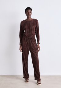 Combinaison bordeaux avec des rayures horizontales, manches longues, taille élastique et jambes évasées. Présente un tissu texturé et une coupe décontractée.