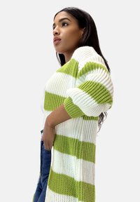 Stickad kofta med djärva vita och limegröna ränder, med lös passform, vida ärmar och ribbad struktur. Bärs över en vit topp.
