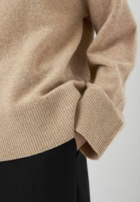 Beżowy sweter wełniany z prążkowanymi mankietami i dołem, prezentujący miękką teksturę. W połączeniu z czarnym ubraniem, podkreślającym kontrast kolorów.