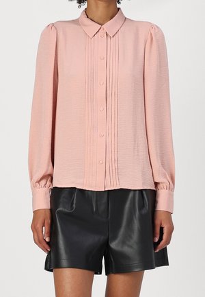 Light pink long-sleeve blouse with front pleats and buttons, paired with black high-waist leather shorts on a standing person.