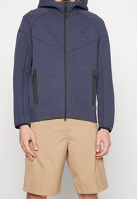 Marinblå zip-up hoodie med huva, två sidofickor och svarta detaljer. Texturerat tyg med en sportig, figurnära design. Bärs över khakifärgade shorts.