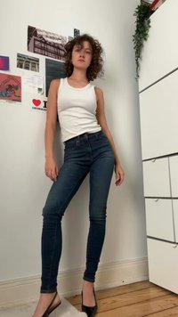 Biela tielko, tmavé modré úzke džínsy a čierne vysoké podpätky. Pozadie obsahuje dekorácie na stene a rastliny. Prírodná drevená podlaha.