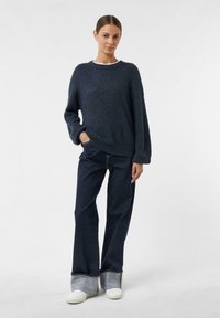 Granatowy sweter z dzianiny z szerokim dekoltem i długimi rękawami, zestawiony z ciemnoniebieskimi spodniami dżinsowymi z podwiniętymi nogawkami i białymi sneakerami.