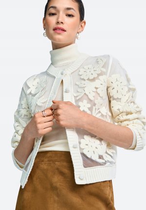Weißer transparenter Cardigan mit floraler Stickerei, geripptem Kragen und Knopfverschluss, getragen über einem cremefarbenen Rollkragenpullover und einem braunen Wildlederrock.