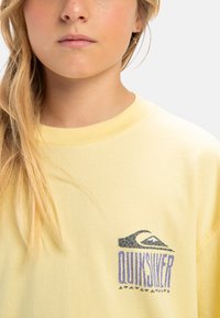 Gele katoenen T-shirt met ronde hals, voorzien van een gebarsten blauwe Quiksilver-logo op de linkerkant van de borst. Zachte textuur, casual ontwerp.