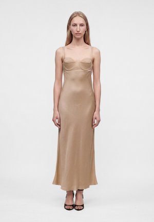 Femme en robe nuisette beige en satin avec bretelles fines et bustier structuré, debout devant un fond blanc uni, portant des talons ouverts noirs.