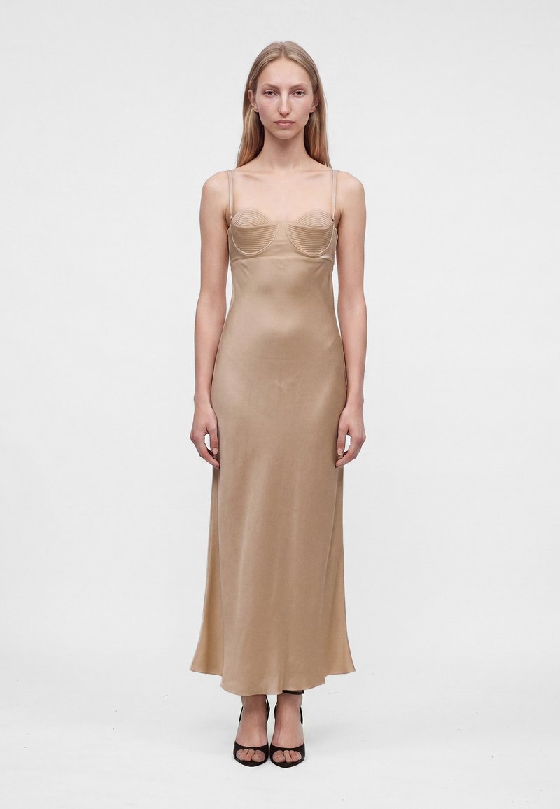 Femme en robe nuisette beige en satin avec bretelles fines et bustier structuré, debout devant un fond blanc uni, portant des talons ouverts noirs.