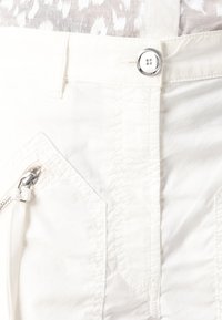 Tessuto bianco con una texture liscia, dettaglio con zip sulla tasca sinistra e un bottone argentato lucido in vita. Le cuciture sono visibili.