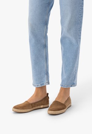 Osoba w jasnoniebieskich dżinsach i brązowych wsuwanych espadrylach stojąca na białym tle.