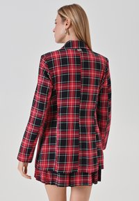 Rot-schwarzer karierter Blazer mit scharfem Kragen, langen Ärmeln und einem Schlitz hinten, der metallische Akzente am Ausschnitt aufweist.