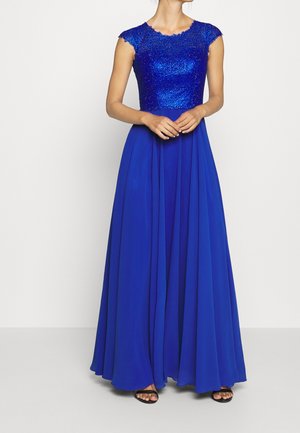 Femme portant une robe longue bleu roi avec un corsage ajusté et scintillant et des manches courtes, debout, mains jointes devant.