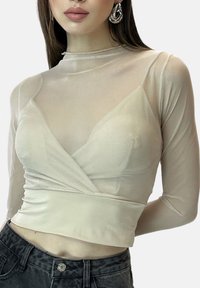 Ljusbeige långärmad topp med genomskinligt överlager och djup V-ringning, utrustad med en figursydd midjeband och slät textur, kombinerad med mörka jeans.