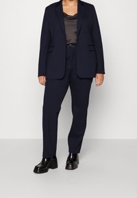 Granatowy, dopasowany garnitur z usztywnioną marynarką i prostymi spodniami, noszony na czarnym satynowym topie. Posiada proste kieszenie i eleganckie buty.