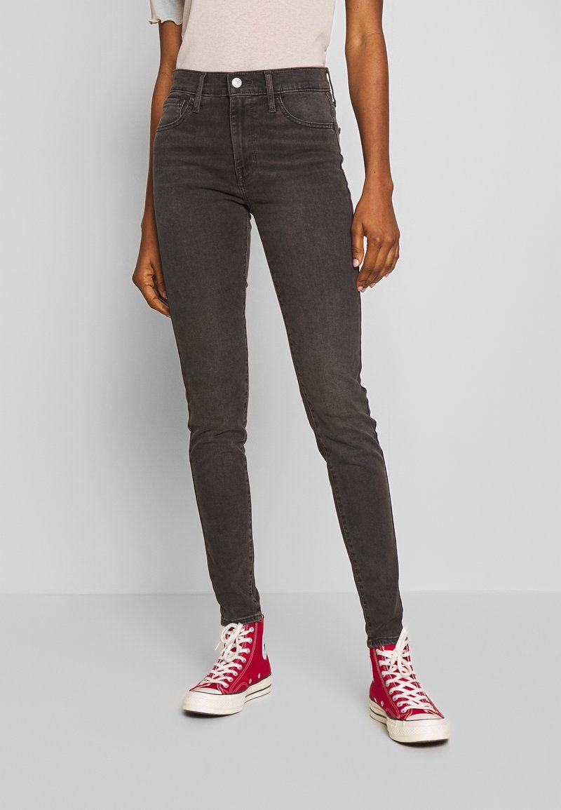 Jean skinny noir en denim à taille haute, avec une fermeture à bouton à l'avant et des poches, associés à des baskets montantes rouges.