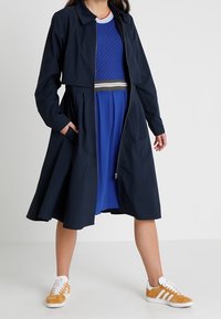 Long manteau bleu marine avec un col et une fermeture éclair, superposé à un pull bleu texturé et une jupe plissée, associé à des baskets blanches et beiges.