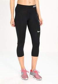 Leggings capri noirs en tissu extensible, dotés d'une taille haute et d'un logo Nike blanc à l'avant. Portés avec des chaussures de sport grises et roses.