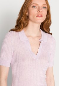 Camisola polo de malha canelada rosa claro, com mangas curtas e um decote em V profundo. O tecido parece suave, com uma textura subtil e um design ajustado.