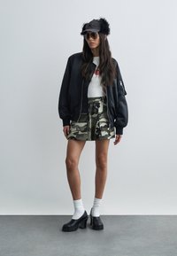 Γυναίκα που φοράει μαύρο μπουφάν τύπου bomber, φούστα παραλλαγής, λευκές κάλτσες με μαύρα χοντρά τακούνια, μαύρο καπέλο με επενδύσεις για τα αυτιά και γυαλιά ηλίου.