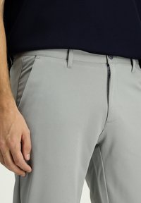 Pantaloni grigi chiari con superficie testurizzata, dotati di cerniera frontale, chiusura a bottone e passanti per cintura. Nessun motivo o accento visibile.