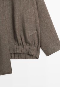 Manche de veste marron texturée avec poignet élastiqué et détail de poche en biais sur un tissu lisse.