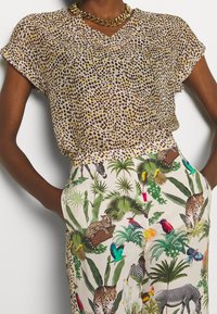 Brązowa bluzka w żółte plamy z dekoltem V, zestawiona z kremowymi spodniami ozdobionymi żywym wzorem dżungli z udomowionymi zwierzętami i roślinnością.