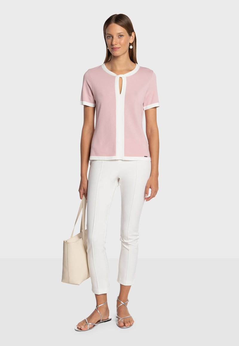 Top rosa chiaro a maniche corte con dettagli bianchi e apertura a goccia, abbinato a pantaloni bianchi a taglio slim e sandali metallici. Borsa tote beige.