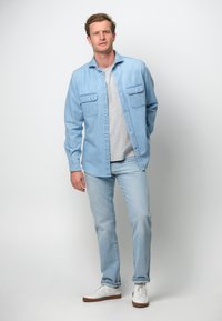 Camisa de mezclilla azul claro con dos bolsillos en el pecho, camiseta gris debajo, jeans azul claro y zapatillas blancas. Conjunto casual y en capas.