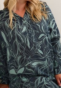 Blouse bleu foncé avec un motif floral vert pâle, dotée d'un col et de boutons, fabriquée à partir d'un tissu doux au toucher lisse.