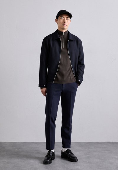 Giacca blu scura con zip bianca, maglione marrone a collo alto, pantaloni navy su misura e scarpe nere con suole spesse, tutti disposti con cura.