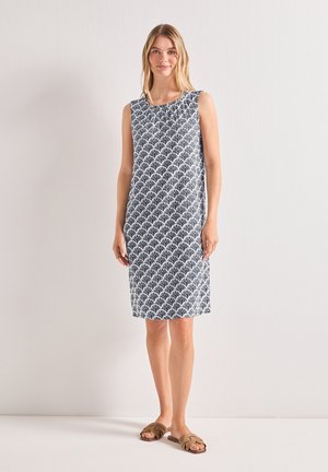 Femme portant une robe sans manches mi-longue au genou avec un motif éventail noir et blanc, debout devant un fond clair uni et des sandales marron.