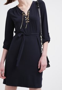 Mörkblå klänning med v-ringning, detaljer med guldkedja, justerbara ärmar och bälte med knytning i midjan. Bärs med en svart handväska.