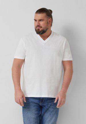 Mann mit Bart und zurückgebundenen Haaren, der ein weißes V-Ausschnitt-T-Shirt und blaue Jeans trägt, vor einem einfachen hellgrauen Hintergrund steht.